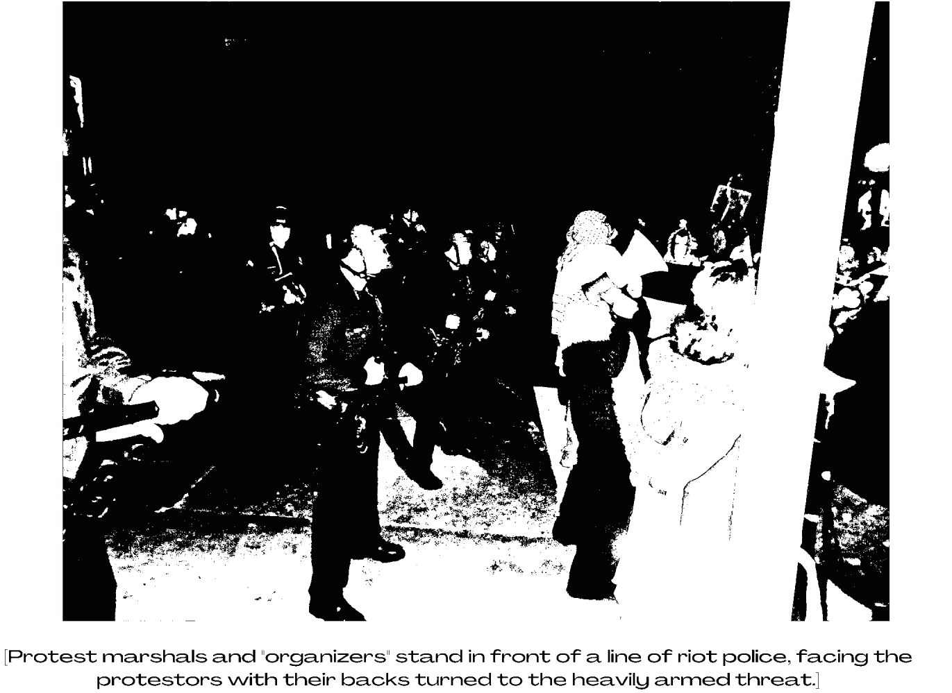 A washed-out, black-and-white photo showing a line of riot police from the side. Just in front of them are people wearing kuffiyehs, one holding a megaphone, facing the same direction as the riot cops. Facing off against this double line is a crowd.